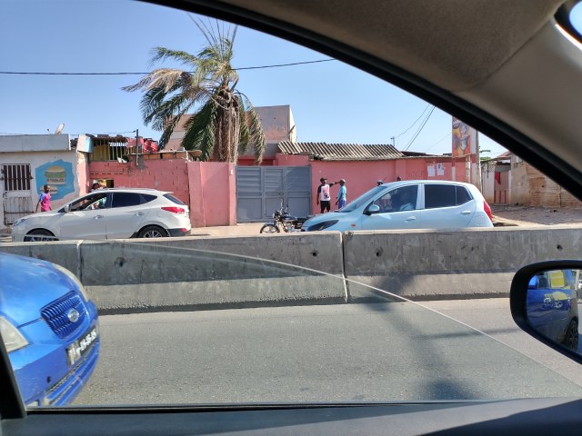 View out of a car window across the road to a pink wall with metal gate.