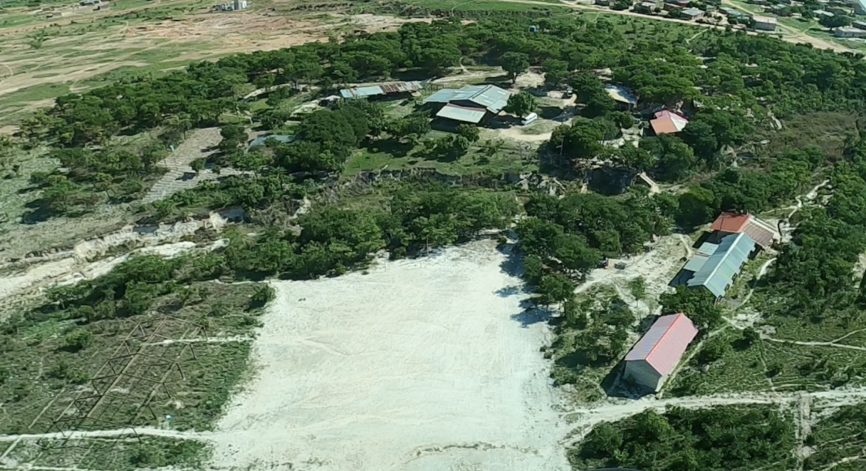 view from an airplane of a plot of land. in the foreground is a large soccer field with a metal frame to the left and three buildings to the right. in the background across from a gully are several structures