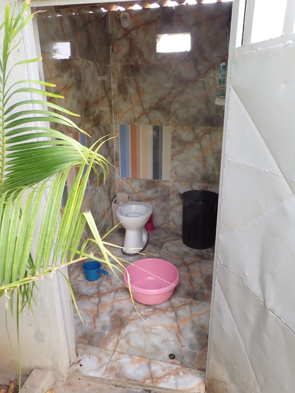 tiled outhouse with toilet bowl, barrel of water, pitcher, shower head, and basin
