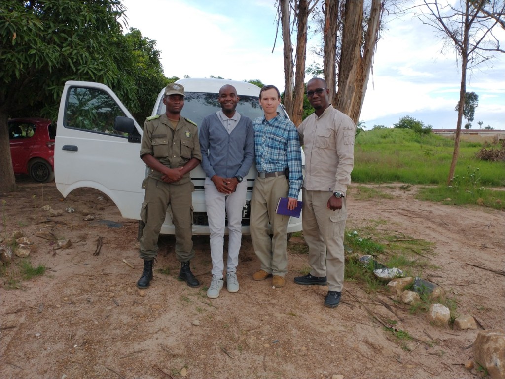 four men, one in uniform, standing in front of a white minivan